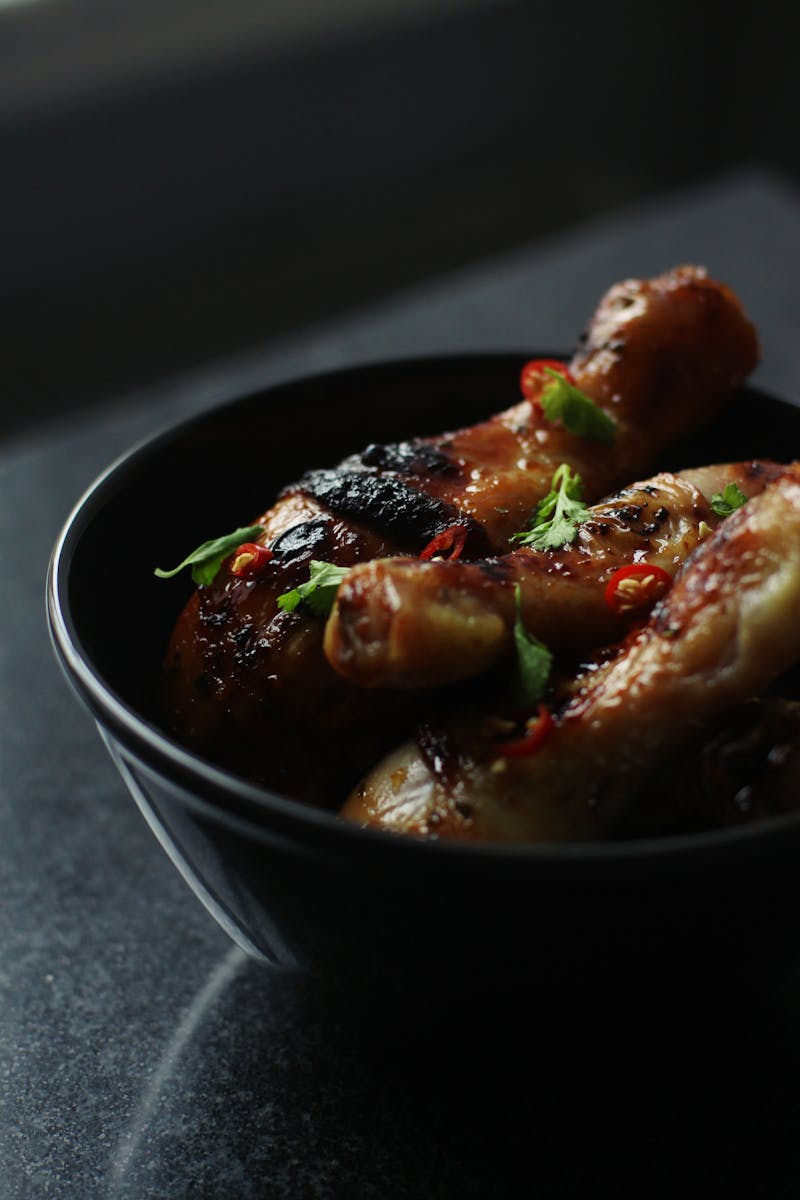 Close-up of roasted chicken legs garnished with parsley and chili, served in a black bowl.