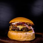 Delicious cheeseburger with melted cheese, onions, and sauce on a wooden board against a dark background.