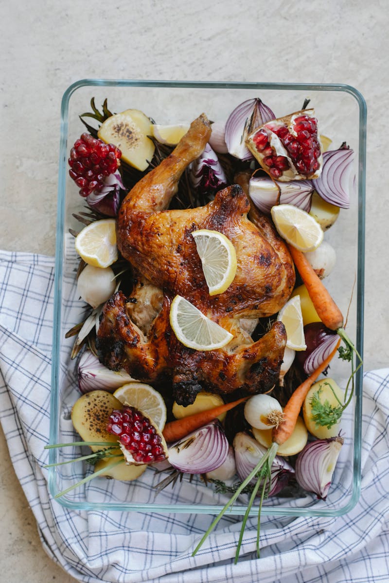 Overhead of tasty grilled whole chicken with lemon and pomegranate placed among onion with carrot served in glass bowl on towel