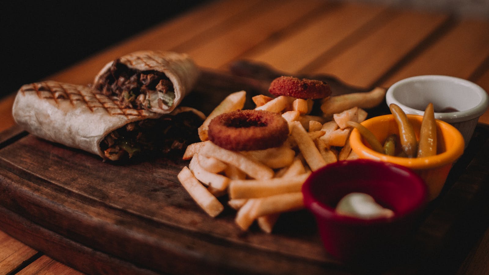 Grilled burrito with fries, onion rings, and sauces served on a wooden board indoors.