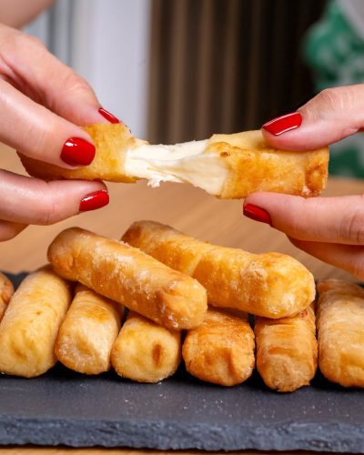 Close-up of hands with red nails pulling apart melted cheese stick, revealing gooey inside.