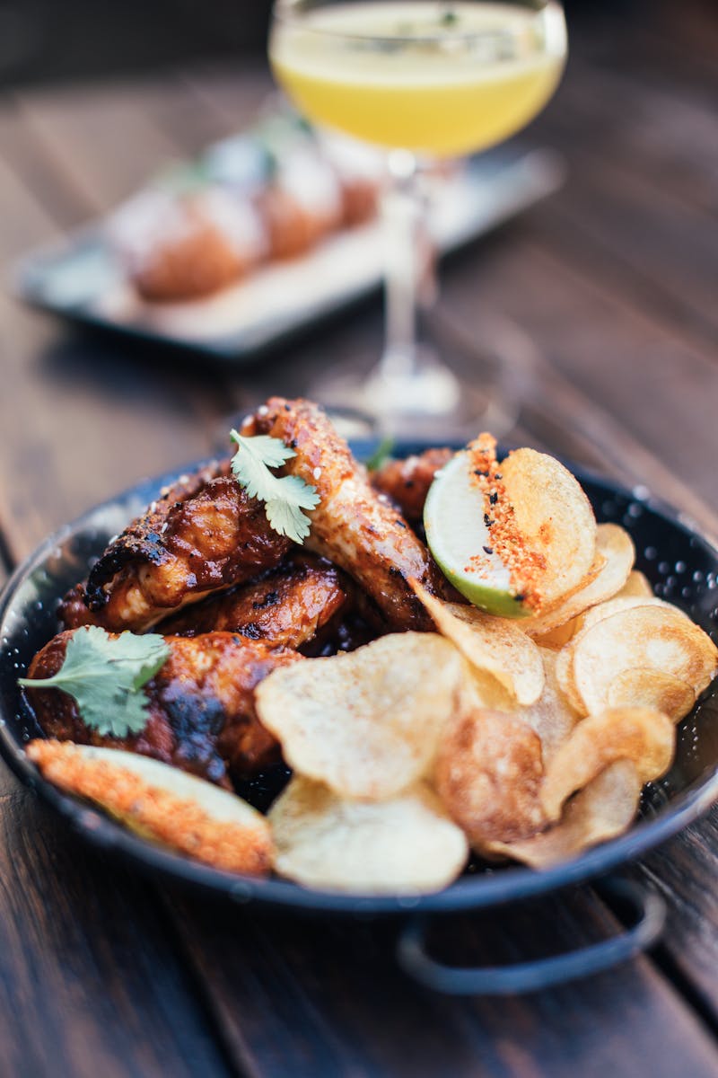 Grilled chicken wings with crisps decorated with herbs placed on wooden table with alcoholic cocktail on blurred background in restaurant