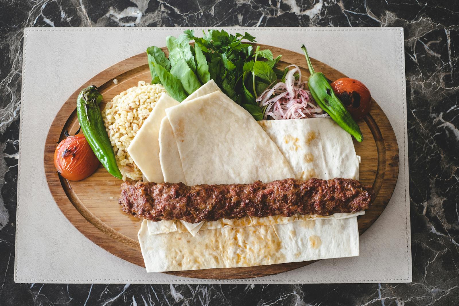 Savory Adana kebab served on wooden plate with fresh greens, tomatoes, and flatbread.