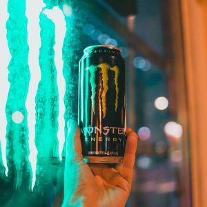 Close-up of hand holding a Monster energy drink against a glowing window at night.