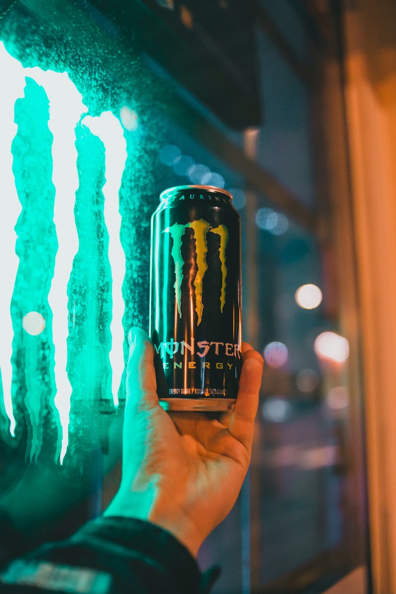 Close-up of hand holding a Monster energy drink against a glowing window at night.